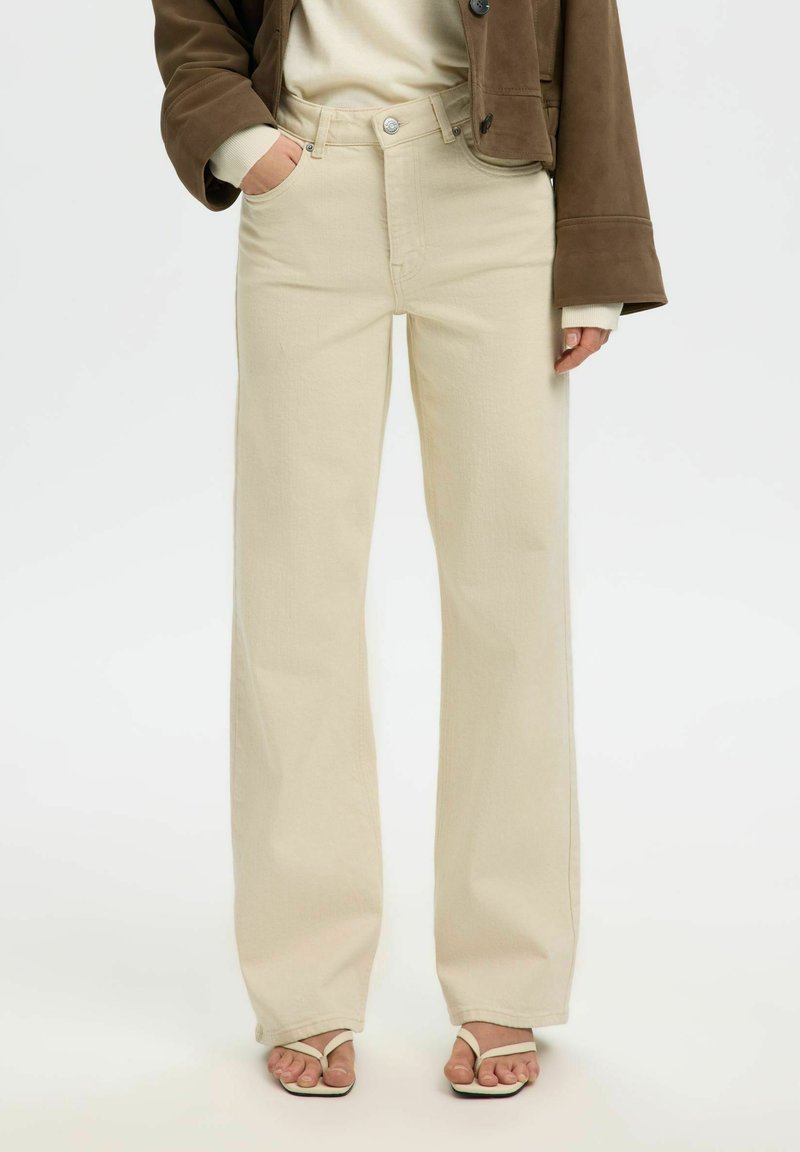 Personne portant un jean large crème, des sandales à talons blanches avec des brides sur les orteils, un haut beige et une veste courte marron.