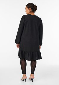 Kvinde med krøllet hår sat op i en knold iført en langærmet sort kjole med flæsekanter, gennemsigtige leggings med prikket mønster og sølvfarvede højhælede sandaler, vendt væk.