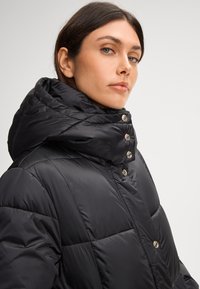 Giubbotto imbottito nero con colletto alto, caratterizzato da dettagli in bottone argentato e design trapuntato. Tessuto liscio, con cappuccio, sopra le spalle del modello.