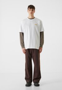 Bijela pamučna majica s malim zlatnim logotipom, složena preko smešane košulje dugih rukava s uzorkom, uparena s smešnim hlačama i crnim cipelama.