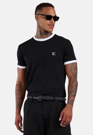 Mann trägt schwarzes T-Shirt mit weißem Rand, schwarzen geflochtenen Gürtel, Sonnenbrille und sichtbare Tätowierungen an Nacken und Armen, stehend vor schlichter Hintergrund.