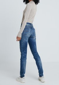 Jeans de mezclilla azul desgastados y de corte ajustado con una cintura de altura media y un leve desvanecimiento. Combinados con una blusa beige de manga larga y zapatillas blancas.