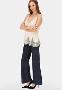 Kremowa bluzka z koronkową lamówką i falowanym dołem, zestawiona z szerokimi ciemnymi dżinsami i czarnymi szpilkami. Gładka faktura, elegancki design.
