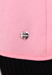 Pink fabric top with a smooth texture, featuring a round silver logo accent near the seam. The lower half shows a black ribbed design.