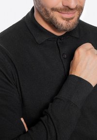 Schwarzes Strick-Polohemd mit gerillter Textur, knöpfbarem Kragen und langen Ärmeln. Nahaufnahme, die die Details und Passform des Shirts hervorhebt.