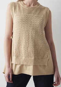 Beige stickad ärmlös topp med en strukturerad, vävd mönster och en ribbad halsringning, lagd över en beige skjorta, tillsammans med mörka jeans.