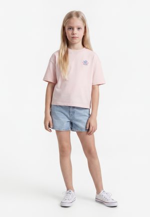 Jeune fille aux longs cheveux blonds portant un t-shirt rose clair, un short en jean et des baskets blanches, debout sur un fond blanc.