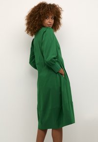 Dlouhá zelená šaty s dlouhými rukávy, volného střihu, s pasovým švem a postranními kapsami. Hladká bavlněná tkanina s matným povrchem.