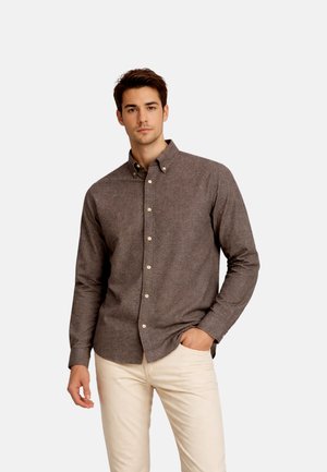 Ung mand iført en brun skjorte med knapper og lange ærmer samt lyse beige bukser, stående med den ene hånd i lommen mod en ensfarvet baggrund.