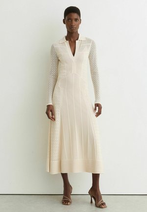 Femme portant une robe en tricot crème à manches longues avec un col et un décolleté en V, associée à des sandales à talons beiges, debout devant un mur uni.