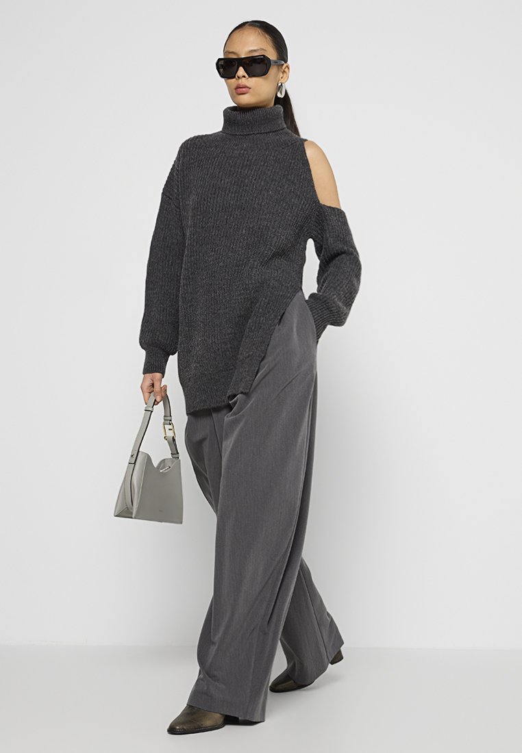 Donna che indossa un maglione dolcevita grigio scuro con spalle scoperte, pantaloni grigi a gamba larga, stivali scuri, occhiali da sole oversize, mentre tiene in mano una borsa a tracolla grigia chiara.