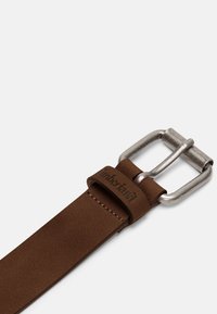 Ceinture en cuir marron avec une boucle métallique argentée. Présente une finition texturée et un logo estampé sur la sangle en cuir.