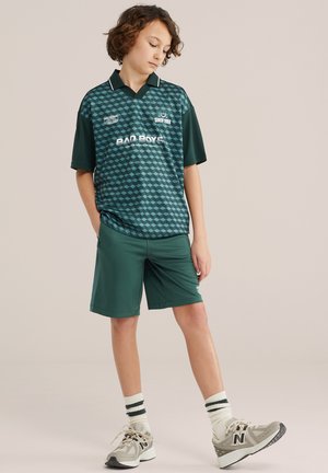 Noor rohelist geomeetrilise mustriga spordisärki ja sobivaid lühikesi pükse kandev noormees, seisab käsi taskus ja vaatab allapoole.