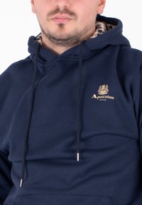 Felpa blu navy con coulisse e punte metalliche. Presenta un piccolo logo dorato sul petto e un motivo a quadri all'interno del cappuccio.