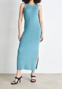 Abito lungo in maglia senza maniche di colore teal con uno spacco laterale, abbinato a sandali neri con tacchi a strappo. Tessuto liscio, silhouette aderente.