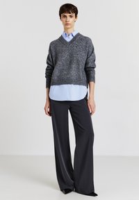Grauer Strickpullover mit V-Ausschnitt, über einem hellblauen Hemd getragen. Schwarze weit geschnittene Hosen runden das Outfit ab. Glatte Texturen und neutrale Farben.