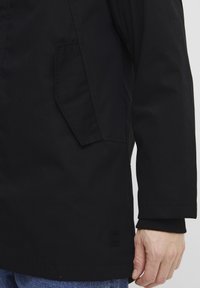 Sort frakke med tekstureret stof, en sidelomme og ribstrikkede manchetter. Designet har en enkel, lige silhuet med minimale accenter.