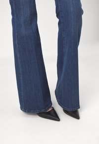 Korkeavyötäröiset tummansiniset leveälahkeiset farkut, joissa on sileä pinta, yhdistettyinä teräväkärkisiin mustiin korkokenkiin. Hemmi on hieman rispaantunut alareunasta.