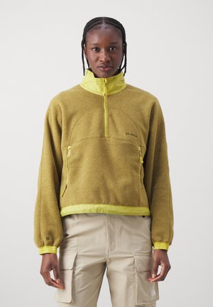 Άτομο που στέκεται φορώντας φλις με μισή φερμουάρ σε μουσταρδί κίτρινο χρώμα, με τσέπες με φερμουάρ, και μπεζ cargo παντελόνι, μπροστά σε απλό φωτεινό φόντο.