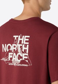 Camiseta de algodón color burdeos con un gran gráfico blanco de montañas y el texto "THE NORTH FACE" con "NEVER STOP EXPLORING" abajo. De mangas cortas.