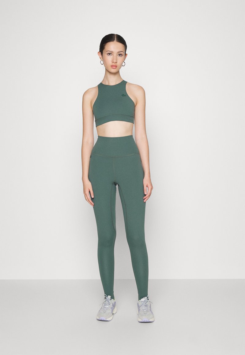 Un crop top de sport vert et un legging taille haute en tissu extensible, associés à des baskets grises. Un design simple avec des détails minimalistes.