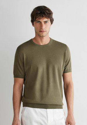 Ung mand med kort brunt hår iført en olivengrøn kortærmet striktrøje og hvide bukser, stående mod en ensfarvet lys baggrund.