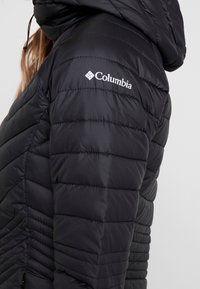 Zwarte gewatteerde jas met capuchon, met wit Columbia-logo op de bovenarm, gedragen door een persoon met lang haar tegen een effen achtergrond.