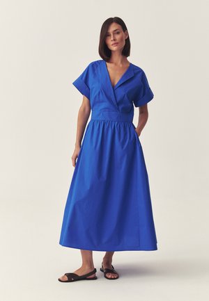 Woman with straight dark hair wearing a short-sleeve cobalt blue midi dress with a V-neck and side pockets, paired with black flat sandals.