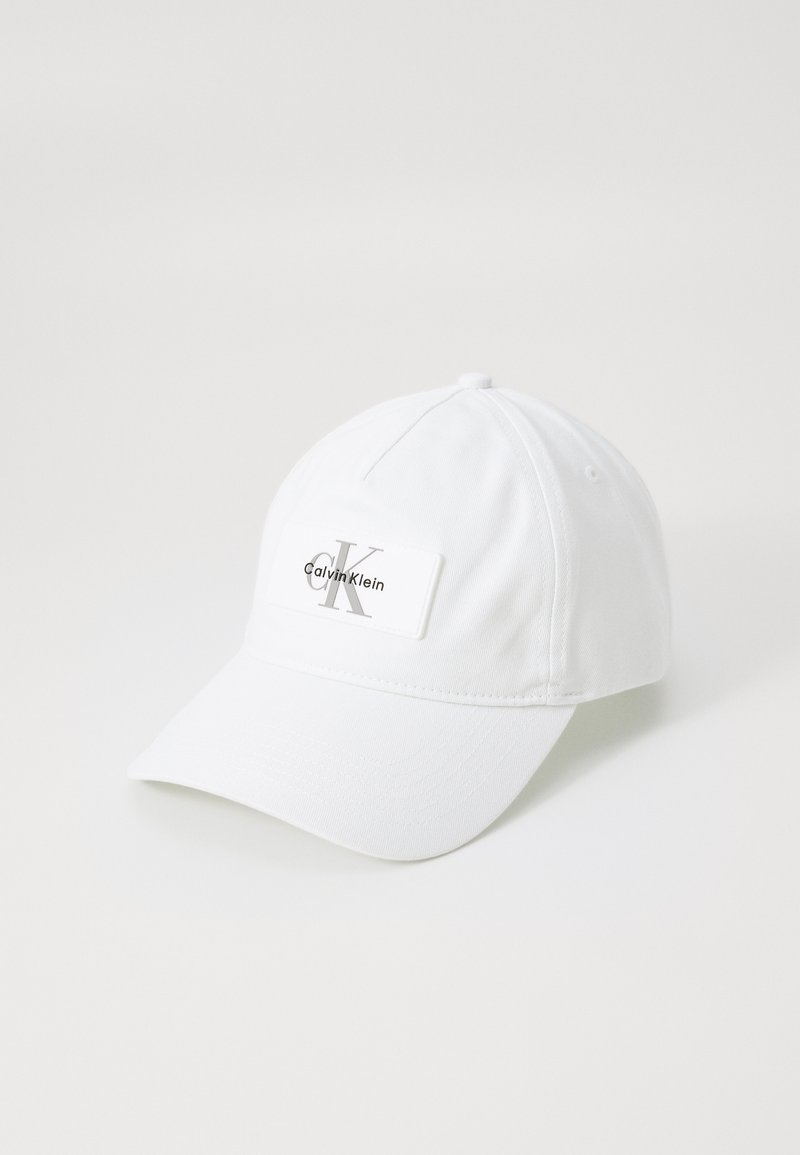 Witte baseballpet met een gebogen rand, stoftextuur, met een rechthoekig label waarop "Calvin Klein" en een gestileerd logo is afgebeeld.