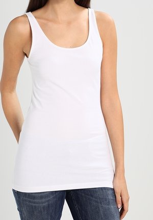 Kvinde iført en tætsiddende hvid ærmeløs tanktop med rund halsudskæring, kombineret med blå jeans.