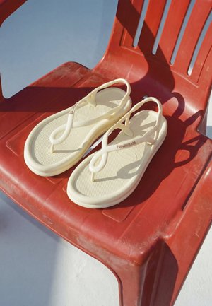 Paar beige Flip-Flops mit Riemen auf einem roten Plastikstuhl, die Schatten auf einen hellen Boden im natürlichen Sonnenlicht werfen.