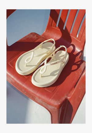 Paar beige Flip-Flops mit Riemen auf einem roten Plastikstuhl, die Schatten auf einen hellen Boden im natürlichen Sonnenlicht werfen.