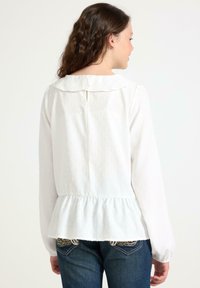 Ung kvinna med lång brunt hår bär en vit, strukturerad långärmad blus med volangkant och mörkblå jeans med broderade bakfickor.