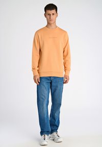 Orange sweatshirt med rund halsringning och ribbade ärmslut. Kombinerad med blå jeans och vita sneakers med flerfärgade snören.