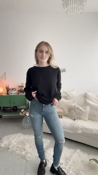 Νεαρή γυναίκα που στέκεται σε ένα σαλόνι φορώντας μαύρο πουλόβερ, σκισμένο ανοιχτό μπλε τζιν και μαύρα παπούτσια, με πράσινη ντουλάπα και καναπέ στο παρασκήνιο.