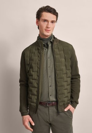 Olive green, quilted puffer jacket with a high collar, paired with a buttoned shirt, brown belt, and matching pants, against a neutral background.
