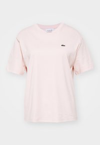 Svetlo roza bombažna majica s kratkimi rokavi, okroglim izrezom in majhnim zelenim logotipom Lacoste na levi strani prsnega koša. Mehak tekstura, sproščen kroj.