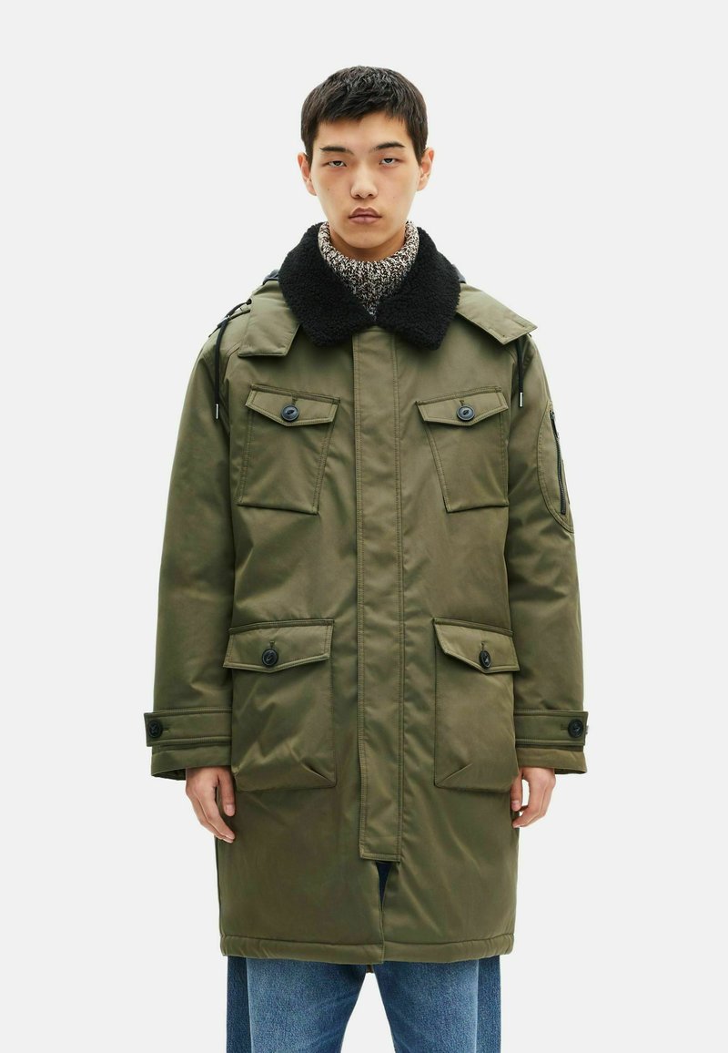 Oliwkowa parka z czarnym futrzanym kołnierzem, licznymi przednimi kieszeniami i zapięciami na guziki, wykonana z gładkiego, wodoodpornego materiału.