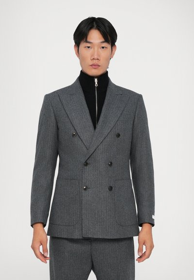 Hombre vestido con un traje gris de doble botonadura con botones negros y un suéter de cuello vuelto negro con cremallera contra un fondo blanco liso.