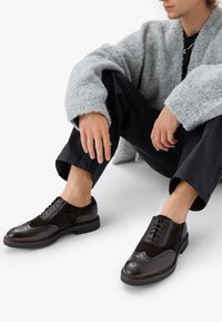 Tummanruskeat oxford-kengät, joissa on brogue-yksityiskohtia, nahkaa ja mokkaa, tasapohjaiset ja pyöreävarvassuunnittelu. Yhdistetty mustiin housuihin.