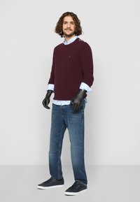 Bordeaux trøje over en lyseblå ternet skjorte, blå jeans og mørke sneakers. Har sorte handsker og et lille logo på trøjen.