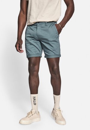 Shorts bleu clair avec ourlets retroussés, assortis à des baskets montantes beiges et des chaussettes beiges avec un logo noir. Coupe et style décontractés.