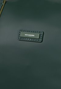 Borsa in materiale sintetico verde scuro con superficie texturizzata, dotata di un'etichetta in pelle cucita con il testo "HEXAGONA" in bianco.