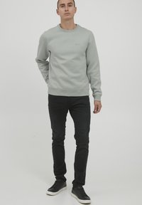 Felpa grigia chiaro con collo a giro, decorata con un sottile logo, abbinata a pantaloni neri slim-fit e sneakers nere. Texture morbida.