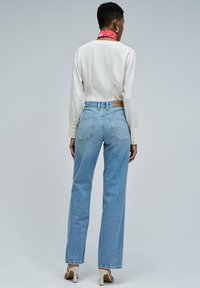Mujer vistiendo jeans de tiro alto de color azul claro, blusa blanca de manga larga, sandalias de tacón blancas y una bufanda de cuello con estampado rojo, de pie mirando hacia atrás.
