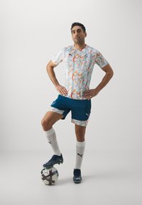 Camiseta de fútbol con un patrón geométrico en blanco, naranja y gris. Combinada con pantalones cortos azules y calcetas blancas hasta la rodilla. De pie sobre un balón de fútbol.