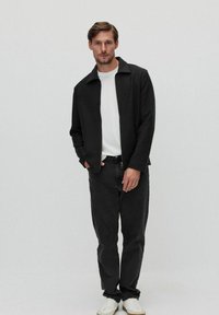 Sort lynlåsjakke lavet af struktureret stof, båret over en hvid skjorte. Sammen med mørke jeans og hvide sneakers mod en lys baggrund.