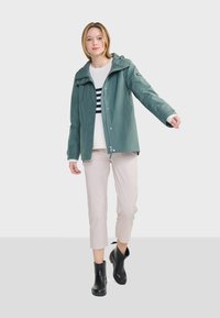 Veste imperméable bleu canard avec fermeture éclair, capuche réglable et poches latérales, associée à un pantalon beige cropped et des bottines noires.