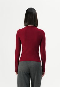 Femme avec des cheveux noirs lisses portant un pull rouge à manches longues en maille côtelée et un pantalon gris cintré, vue de dos sur un fond uni.