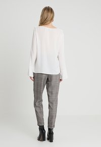 Blusa bianca a maniche lunghe con polsini svasati, abbinata a pantaloni a sigaretta grigi a quadri con accenti rossi e neri, indossata con stivali neri con tacco a blocco.